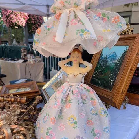 A vintage figurine lamp with a floral skirt and lampshade sits on a table at an outdoor market.