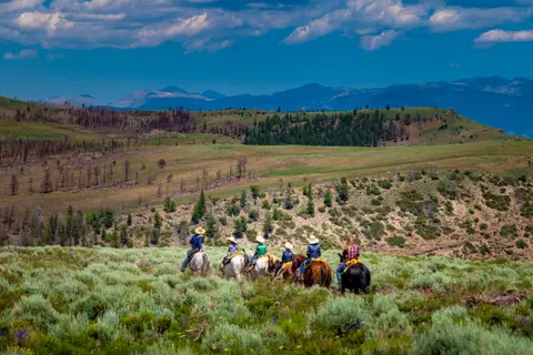 A group of people horseback riding through a grassy landscape with mountains in the background.