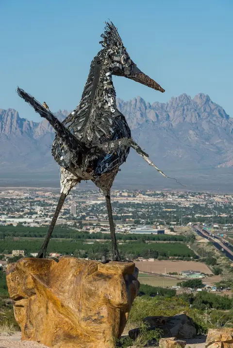 A large roadrunner sculpture made of recycled metal and debris stands overlooking a valley and mountains.
