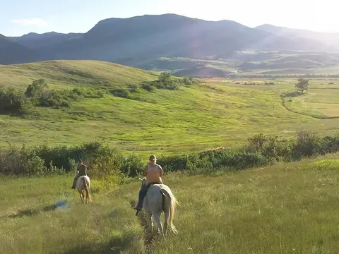 Two people ride horses through a grassy valley with mountains in the background.