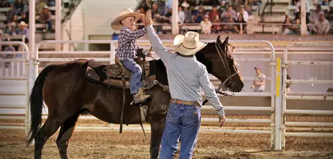 A young boy on a horse high-fives a man in an arena.