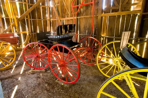 An interior shot shows old carriages with red and yellow wheels inside a rustic wooden barn.