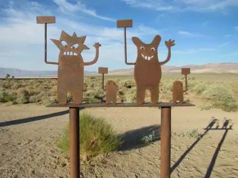 Metal sculpture of cartoon figures holding signs in a desert landscape.