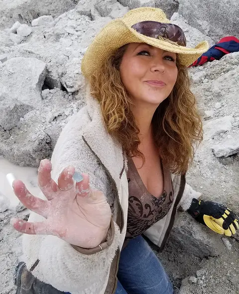 A woman wearing a cowboy hat holds up a small mineral specimen outdoors.