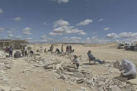 People dig and break stones at an outdoor site under a cloudy sky.