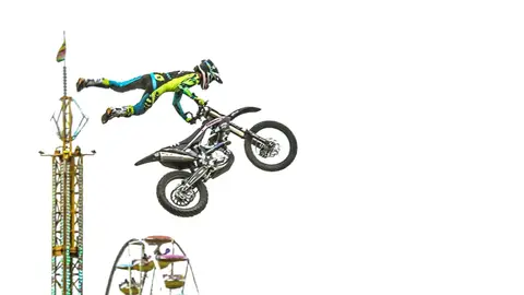 A motocross rider performs a stunt in midair next to fairground rides.