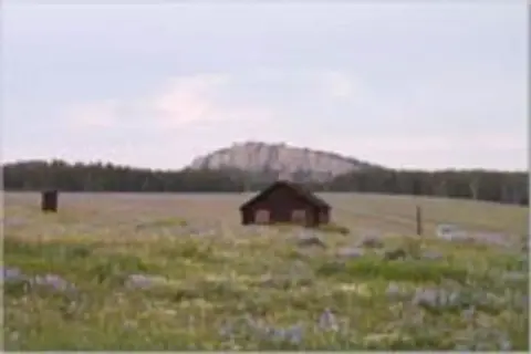 A small cabin in a grassy field with a rocky hill in the background.