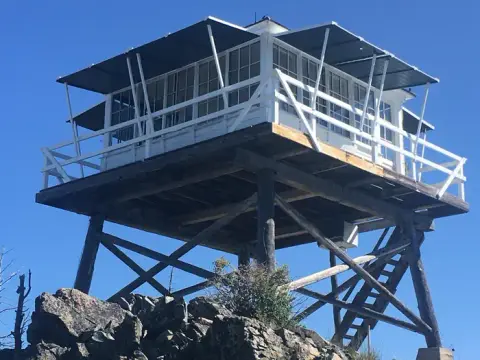 A tall wooden fire lookout tower stands on a rocky summit against a clear blue sky.