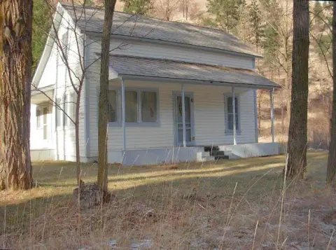 A small white wooden house with a porch stands among trees and dry grass.