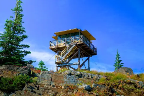 A fire lookout tower stands on a rocky mountain ridge under a blue sky.