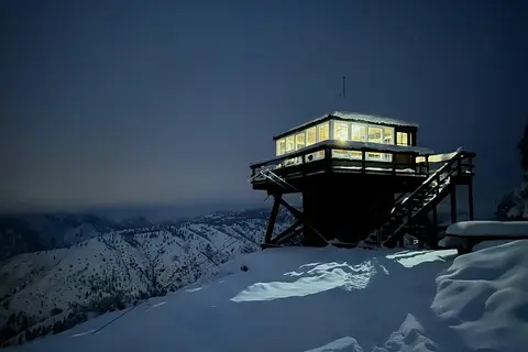 A lighted fire lookout tower sits atop a snowy mountain peak at night.