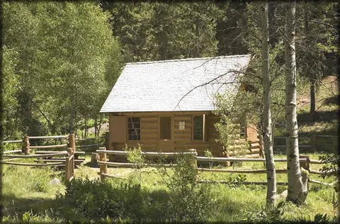 A rustic log cabin stands in a wooded clearing surrounded by a split-rail fence.