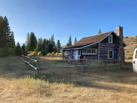 A log cabin stands in a grassy clearing with a wooden fence and trees in the background.