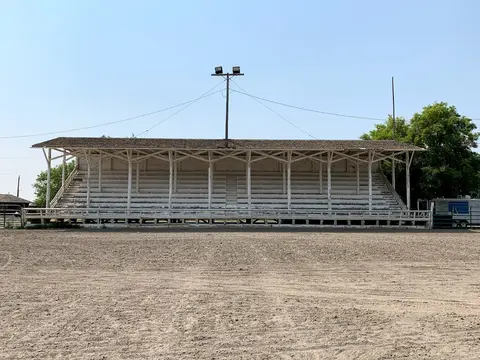 A rustic white wooden grandstand stands in front of a dirt field under a clear blue sky.