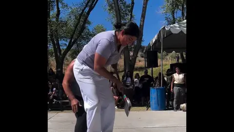 A person swings an axe outdoors in front of a crowd and tents.