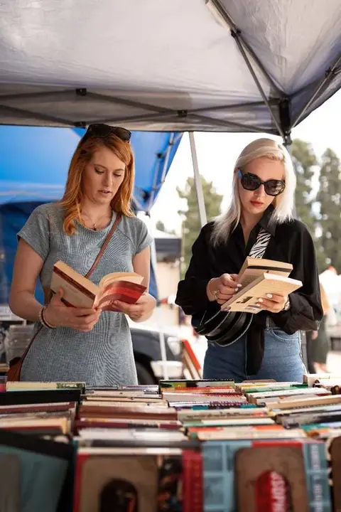 Two women browse books at an outdoor market stall under a tent.