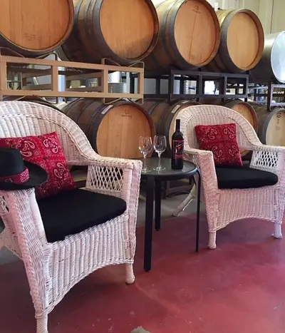 Two white wicker chairs and a table are set in front of wine barrels inside a winery.
