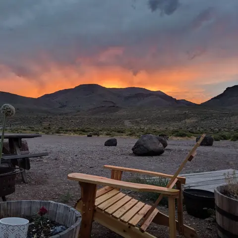 A wooden chair sits outdoors facing a sunset over rolling mountains and a desert landscape.