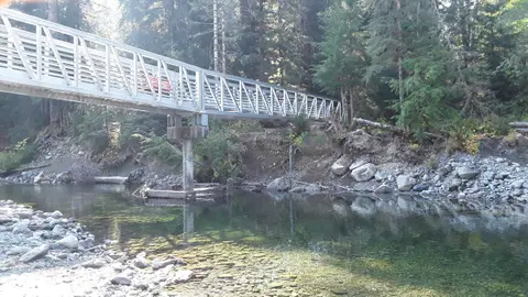 A metal pedestrian bridge crosses a clear river surrounded by trees.