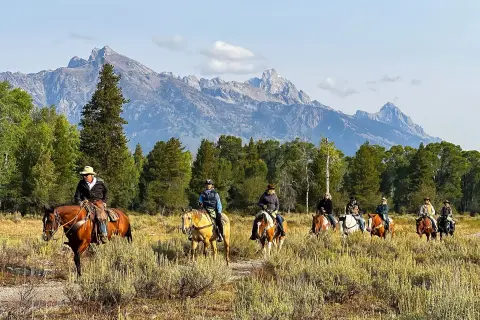 A group of people horseback riding through a grassy field with mountains in the background.