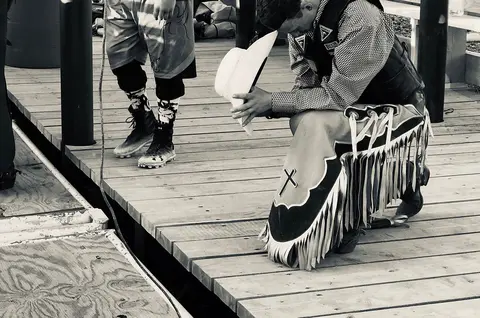 A cowboy kneeling on a wooden dock holding a hat.
