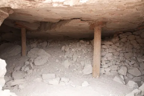 A low-ceilinged underground space supported by wooden posts with loose rocks and stones.