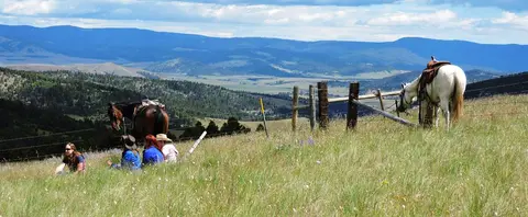 People and horses resting on a grassy hill with a view of mountains in the background.