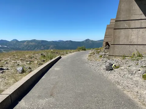 A paved walkway leads past a large concrete structure with distant mountains and a blue sky in the background.