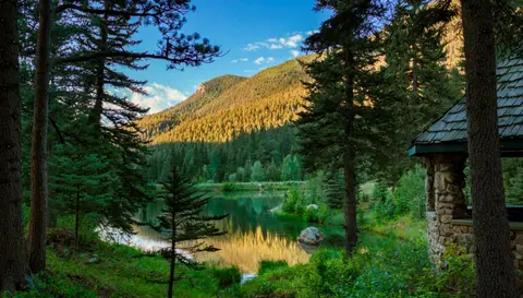 A scenic view of a mountain lake surrounded by pine trees with a small stone structure on the right.