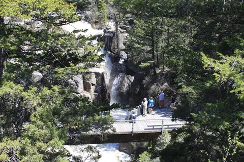 People stand on a bridge looking at a waterfall flowing through a rocky gorge.
