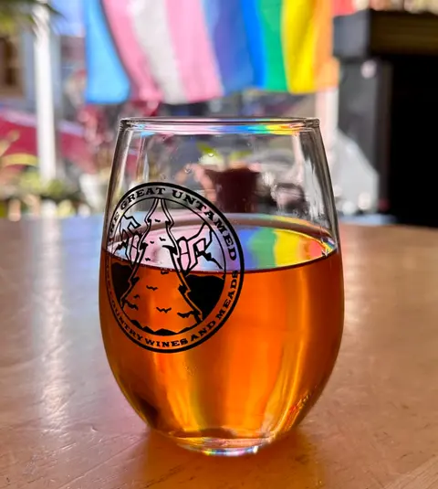 A glass of amber drink sits on a wooden table with a logo printed on it.