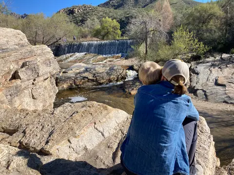 Two people sit on rocks overlooking a small waterfall in a desert landscape.