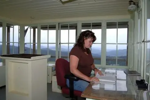 A woman sits at a desk inside a fire lookout tower with windows looking out over mountains.