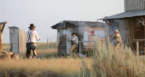 Men engaged in a gunfight outdoors near rustic wooden buildings.