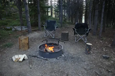 A campfire burns in a metal ring at a campsite in the woods with folding chairs and logs.