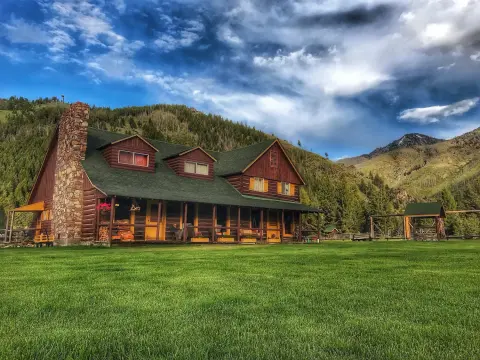 A large wooden cabin with a stone chimney sits in a grassy clearing against a backdrop of mountains and a cloudy sky.