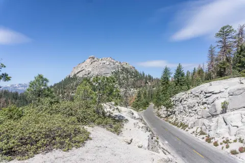 A scenic paved road winding through a mountainous landscape with rocky peaks and pine trees.