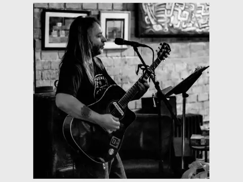 A musician plays an acoustic guitar and sings into a microphone indoors.
