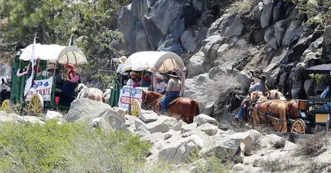 A line of covered wagons and horses travels along a rocky path next to a steep cliff.