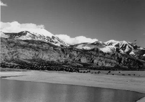 A black and white landscape showing a body of water in the foreground and snowy mountains in the background.