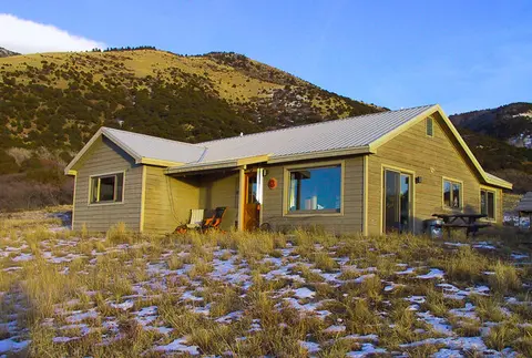A one-story wooden cabin stands in a grassy field in front of a mountain.