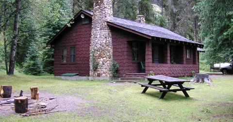 A rustic brown cabin in a wooded area with a stone chimney and picnic table.