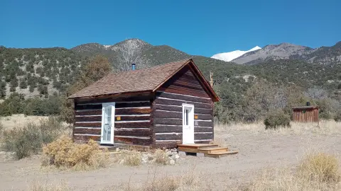 A rustic wooden cabin sits in a dry landscape with mountains in the background.