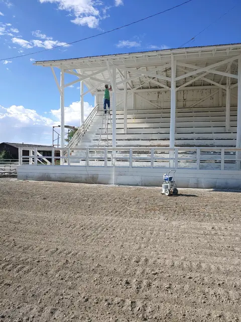 A person paints a white wooden grandstand structure outdoors.
