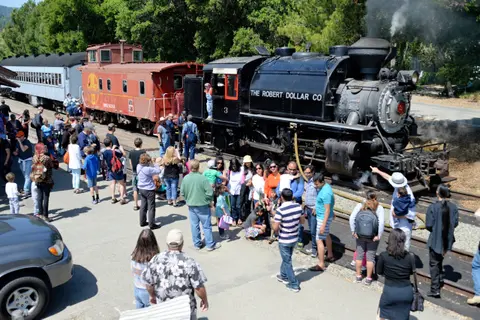 A crowd of people gathered around a historic steam train and cars.