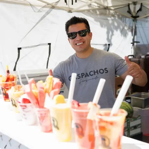 A smiling vendor gives a thumbs up behind a counter of fruit drinks.