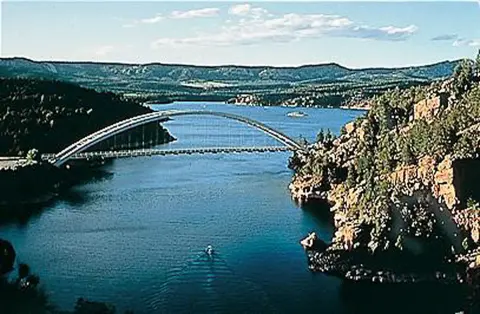 A large arch bridge spans a body of water surrounded by wooded hills.
