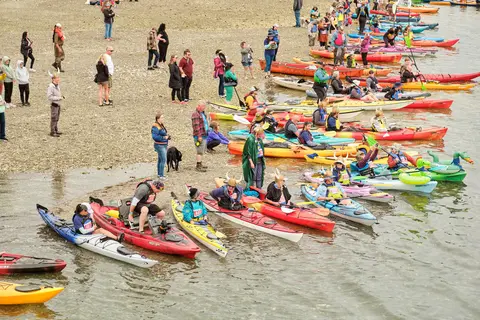 An elevated photograph shows many people with kayaks gathered along a shoreline and water.