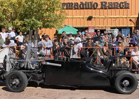 A custom black hot rod vehicle is parked in front of a crowd and a building.