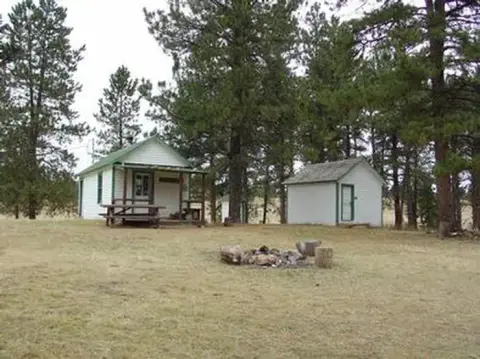 Two small white buildings sit in a clearing surrounded by tall pine trees.